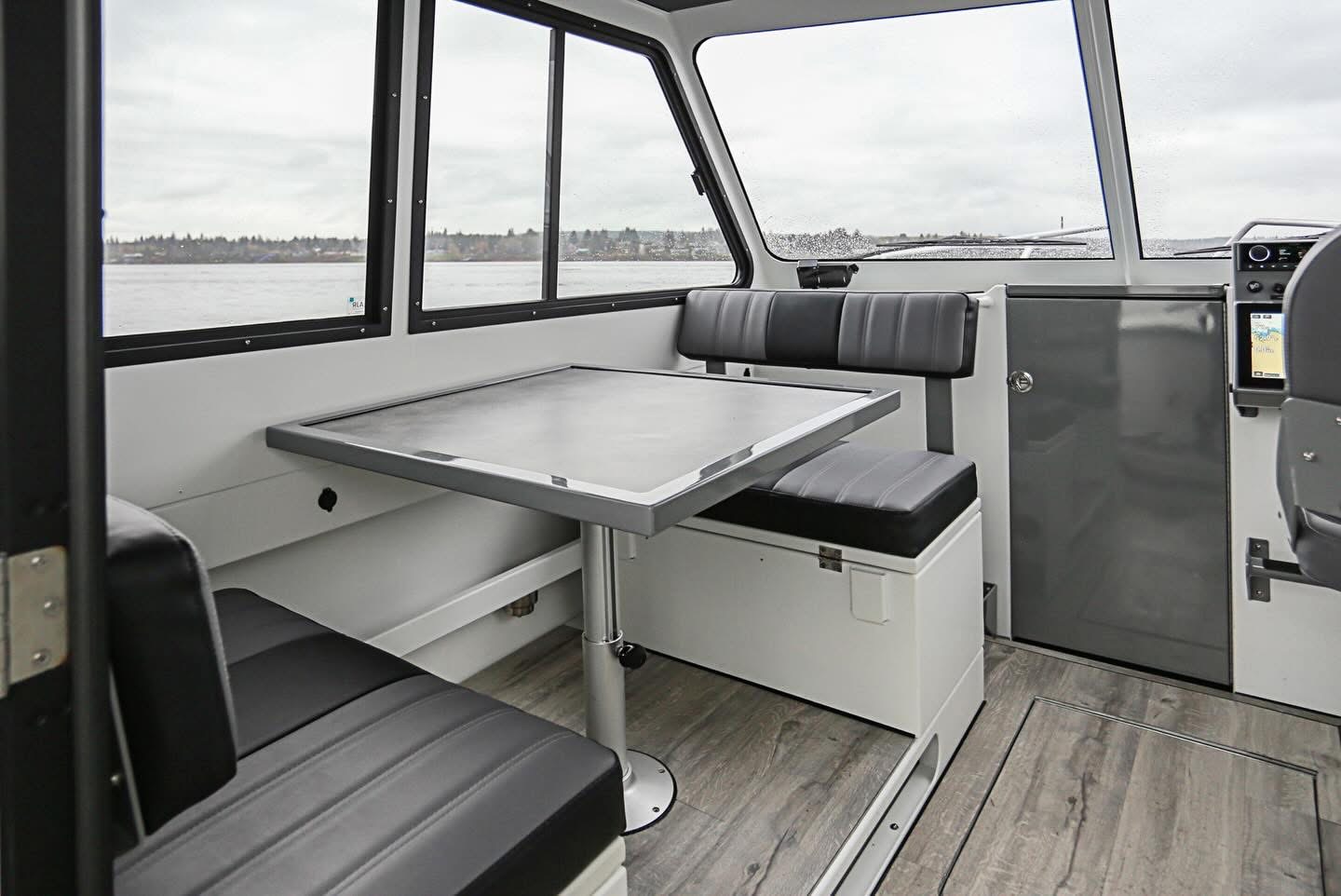 Kwigwis Adventures boat interior cabin featuring comfortable black ribbed seating benches, a table with metal frame, and multiple large windows showcasing calm water and forested shoreline beyond.